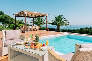 Un espace aménagé au bord de la piscine, avec une table dressée pour le petit-déjeuner, une pergola et une vue sur la mer.