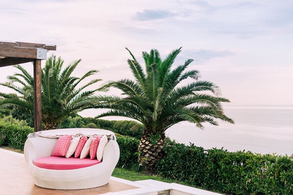A white wicker lounge chair with pink cushions, positioned outdoors with a view of the sea and palm trees.