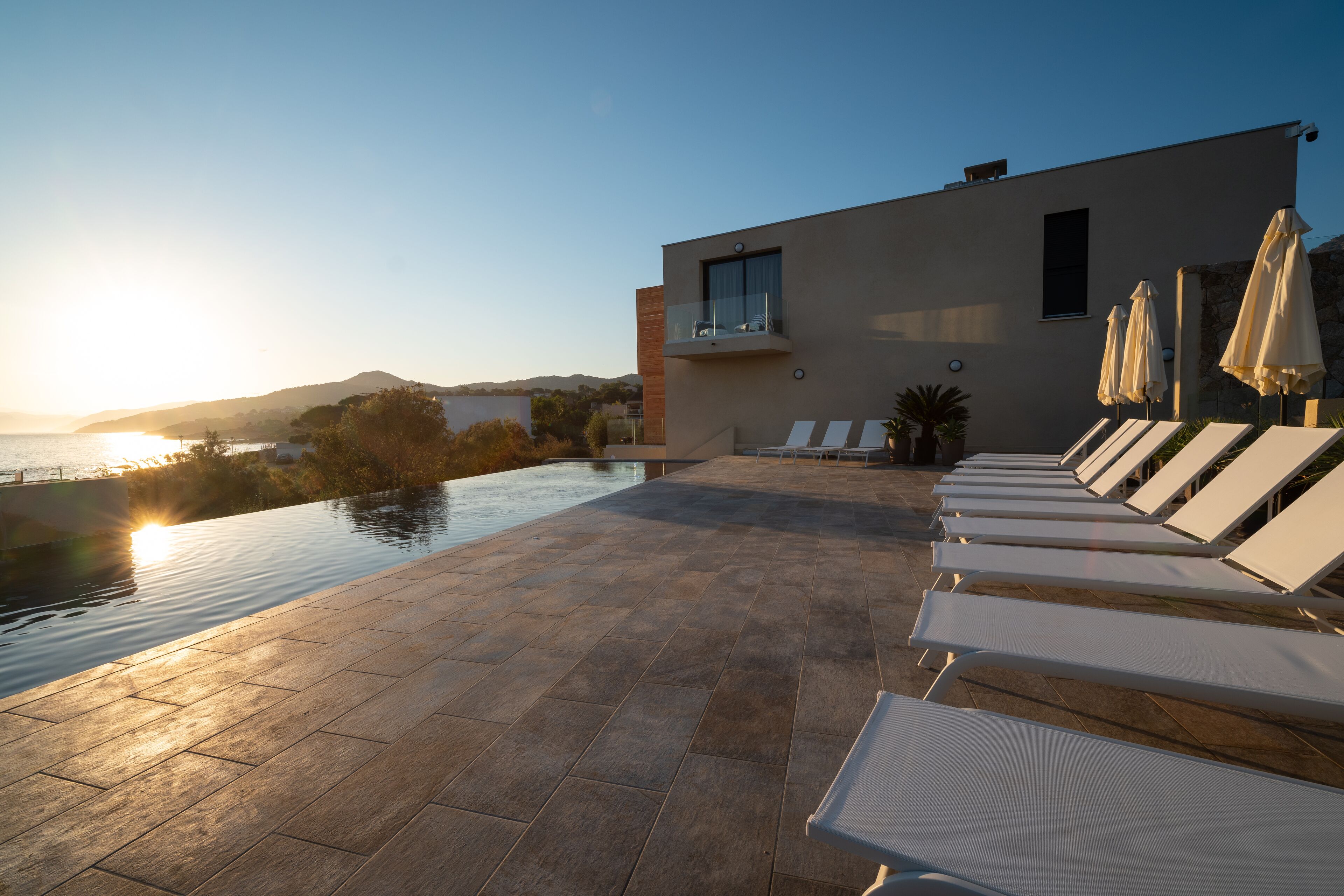 Ein Poolbereich im modernen Stil mit Liegestühlen und Sonnenschirmen, mit Blick auf ein Gewässer und eine Berglandschaft.