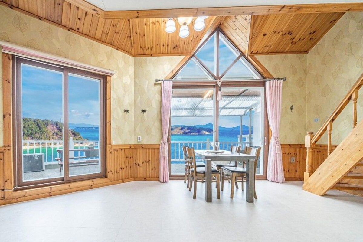 A room with a large window, a dining table with chairs, and a wooden staircase.