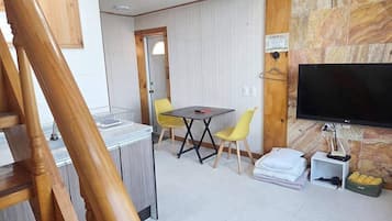 Una habitación con una escalera de madera, una mesa de comedor con sillas amarillas, un televisor de pantalla plana montado en una pared de piedra y un pequeño estante blanco con una lámpara.