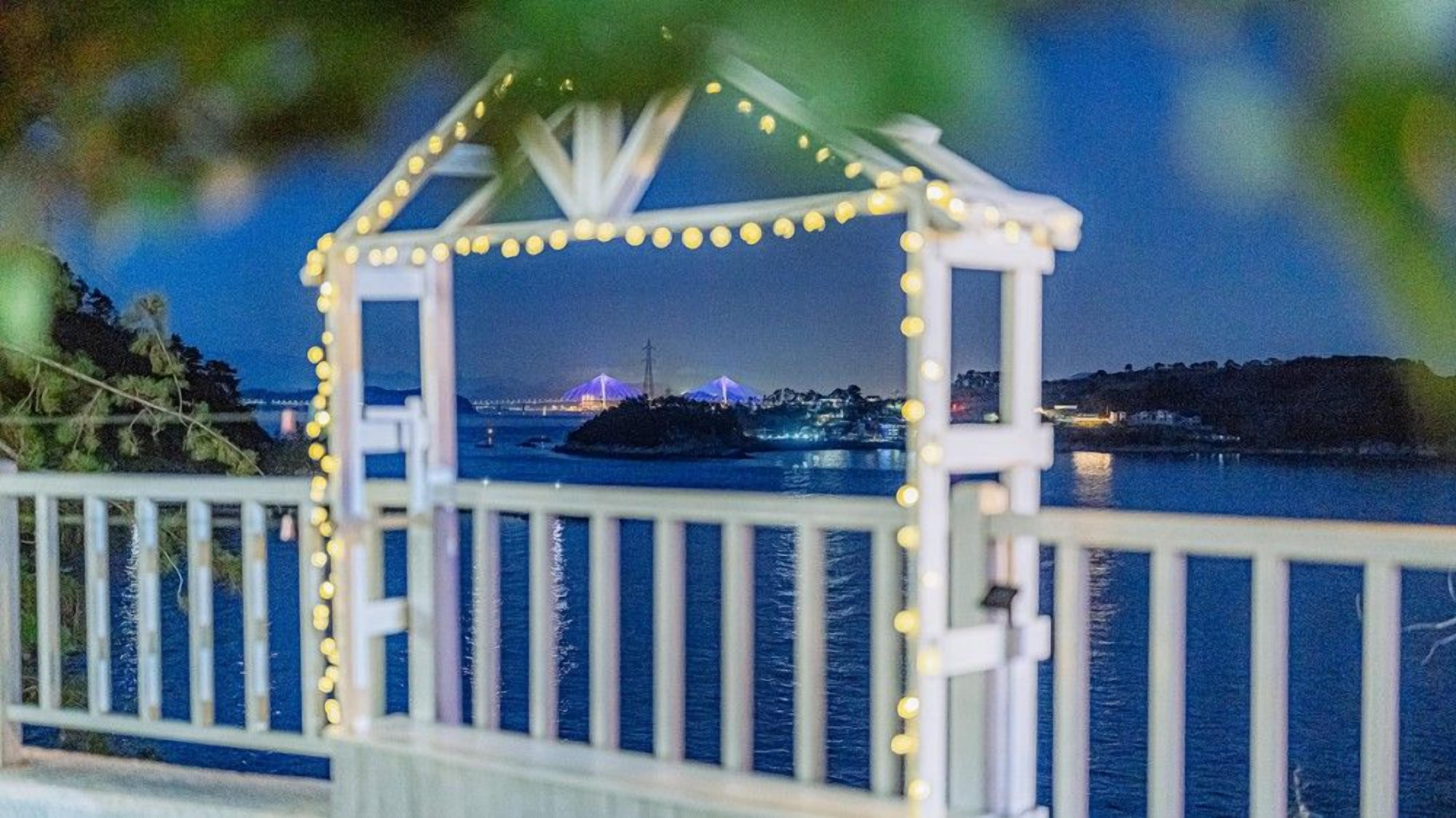A white gazebo with string lights, overlooking a body of water with a distant illuminated structure.