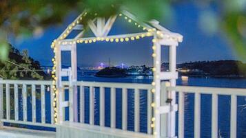 A white gazebo with string lights, overlooking a body of water with a distant illuminated structure.