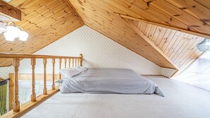 A bedroom with a sloped ceiling, wooden beams, a bed with white bedding, and a wooden railing. - White O Pension (Geoje)