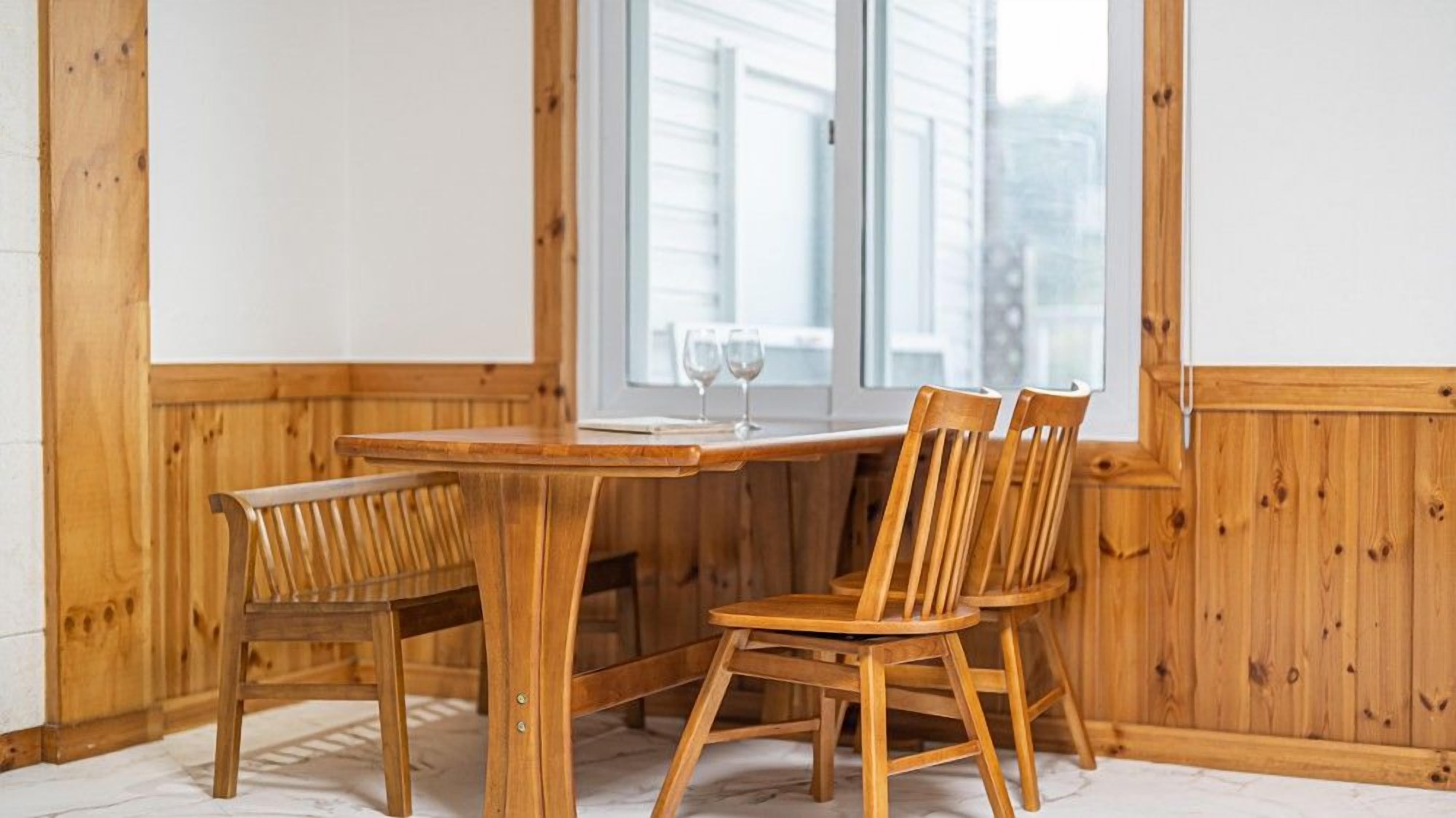 A wooden dining table with two chairs, a window with blinds, and a view of a building outside.