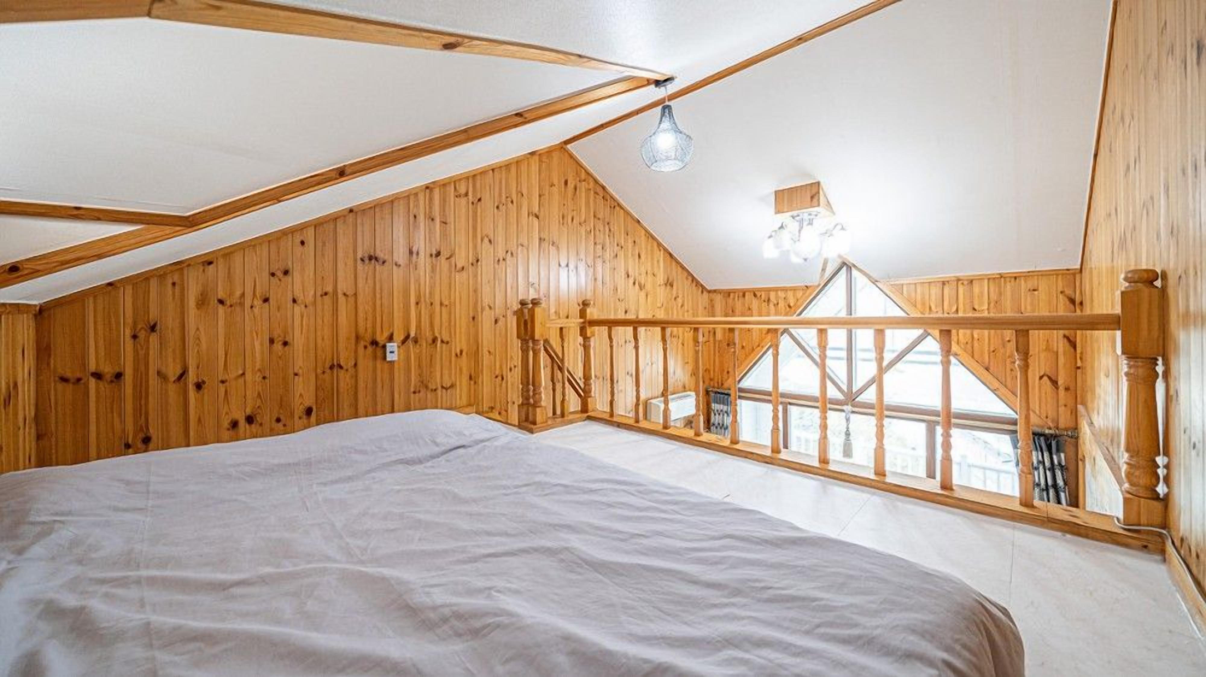 Ein Schlafzimmer mit schrägverdeckter Decke, Holzwänden, einem Bett, einem Fenster und einer hängenden Leuchte.