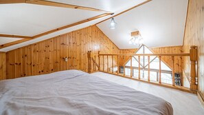 A bedroom with a sloped ceiling, wooden walls, a bed, a window, and a hanging light fixture. - White O Pension (Geoje)