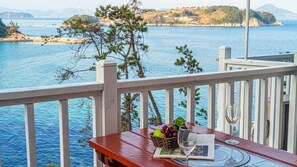 A wooden table with a basket of fruit, a glass of water, and a book on a balcony overlooking a body of water with distant land and a bridge. - White O Pension (Geoje)