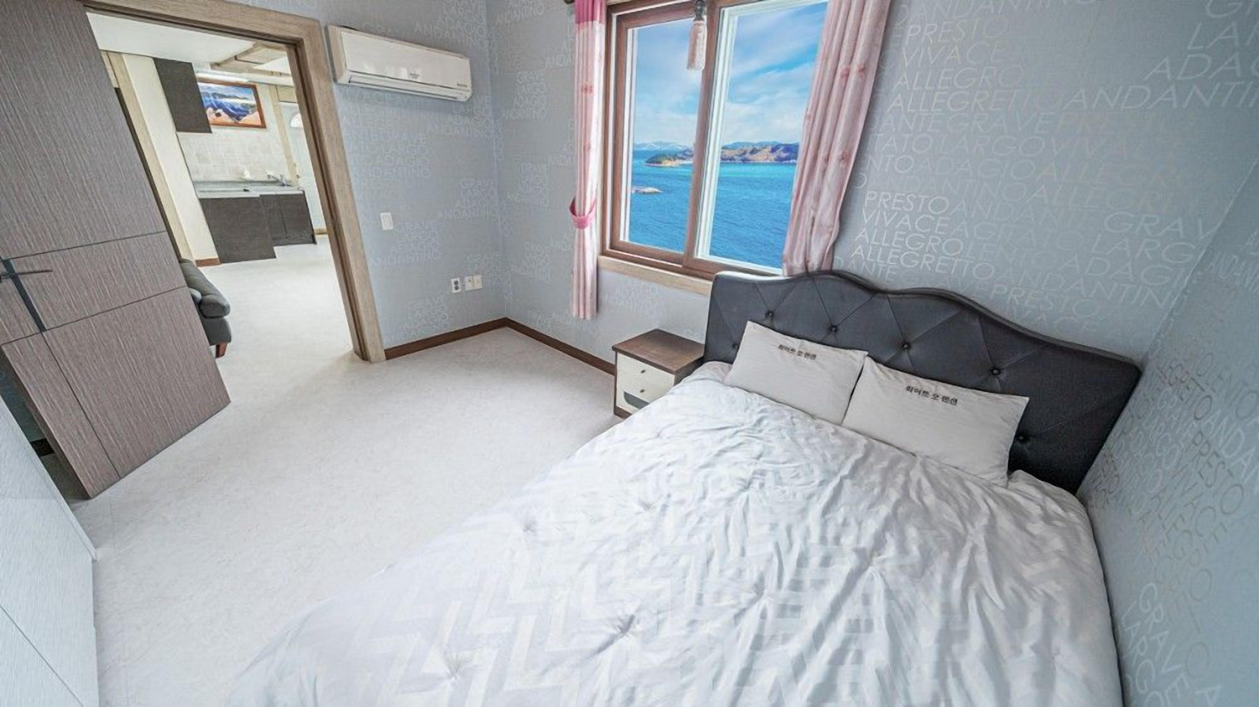 Quarto com cama, mesa de cabeceira, janela com vista para o mar e porta que dá para o banheiro.