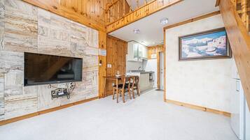 Ein gemütliches Blockhaus-Interieur mit Steinwand, Holzboden und einem an der Wand befestigten Fernseher. Es gibt einen Essbereich mit Stühlen und eine Küche mit Schränken.
