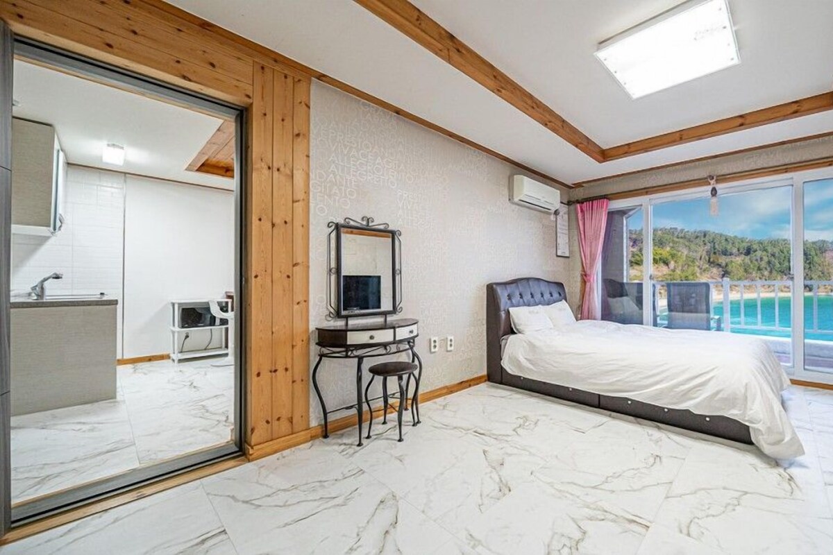 A bedroom with a bed, a desk, a mirror, and a view of a beach.
