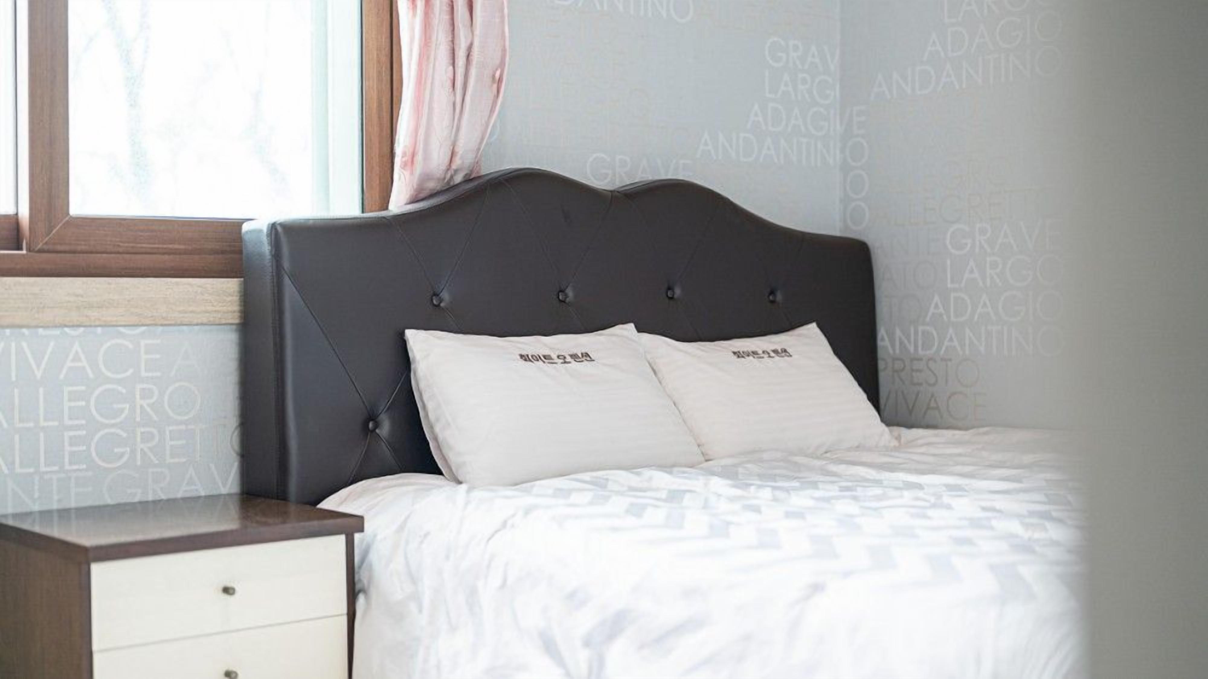 Una cama bien hecha con un cabecero oscuro y dos almohadas, una mesita de noche con cajón y una ventana con cortina.