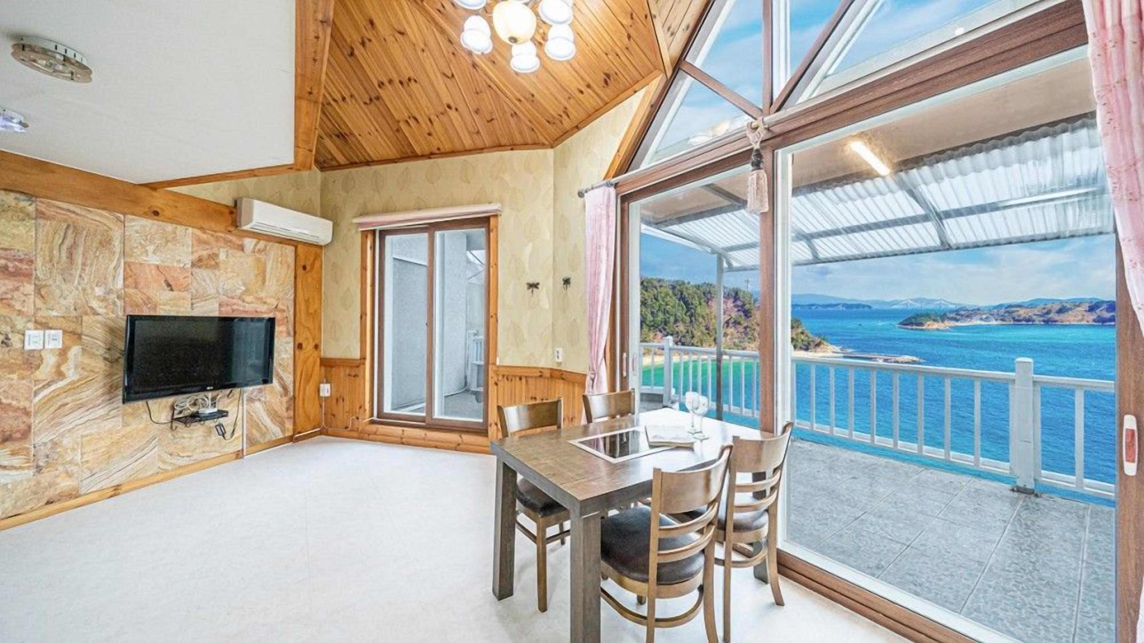 Habitación con un ventanal grande que ofrece vistas panorámicas de la costa y un puente, un comedor con mesa y sillas, y un televisor instalado en una pared de piedra.