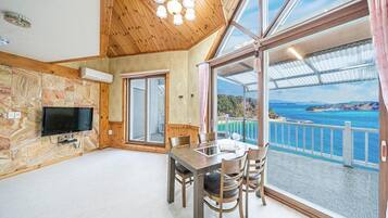 Habitación con un ventanal grande que ofrece vistas panorámicas de la costa y un puente, un comedor con mesa y sillas, y un televisor instalado en una pared de piedra.