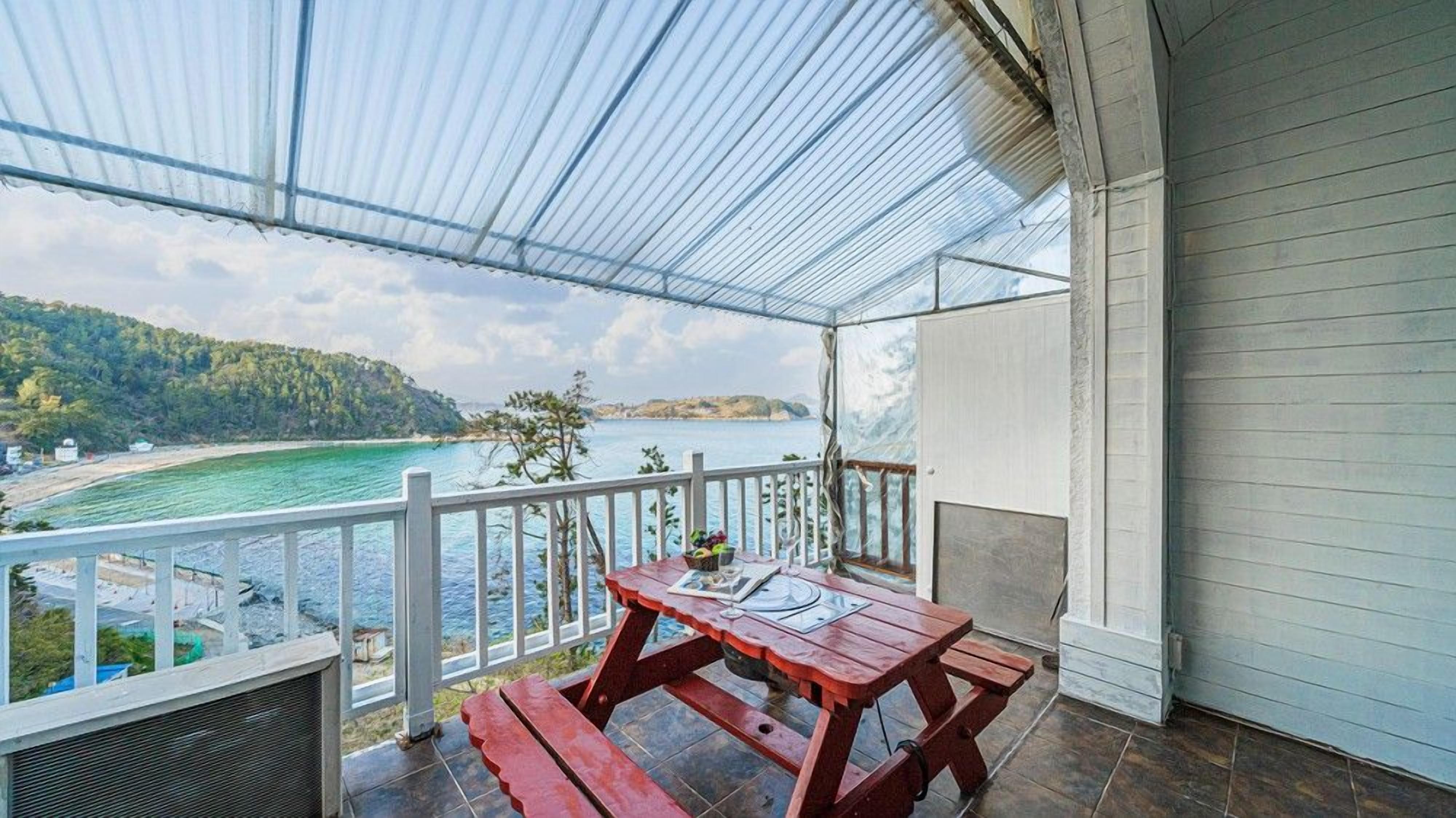 Un balcón techado con una mesa y bancos, con vista a una zona costera de aguas azules y una costa distante.