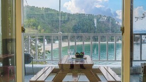 A wooden table with a bowl of fruit and two glasses on a balcony with a white railing, overlooking a beach and forested hills. - White O Pension (Geoje)