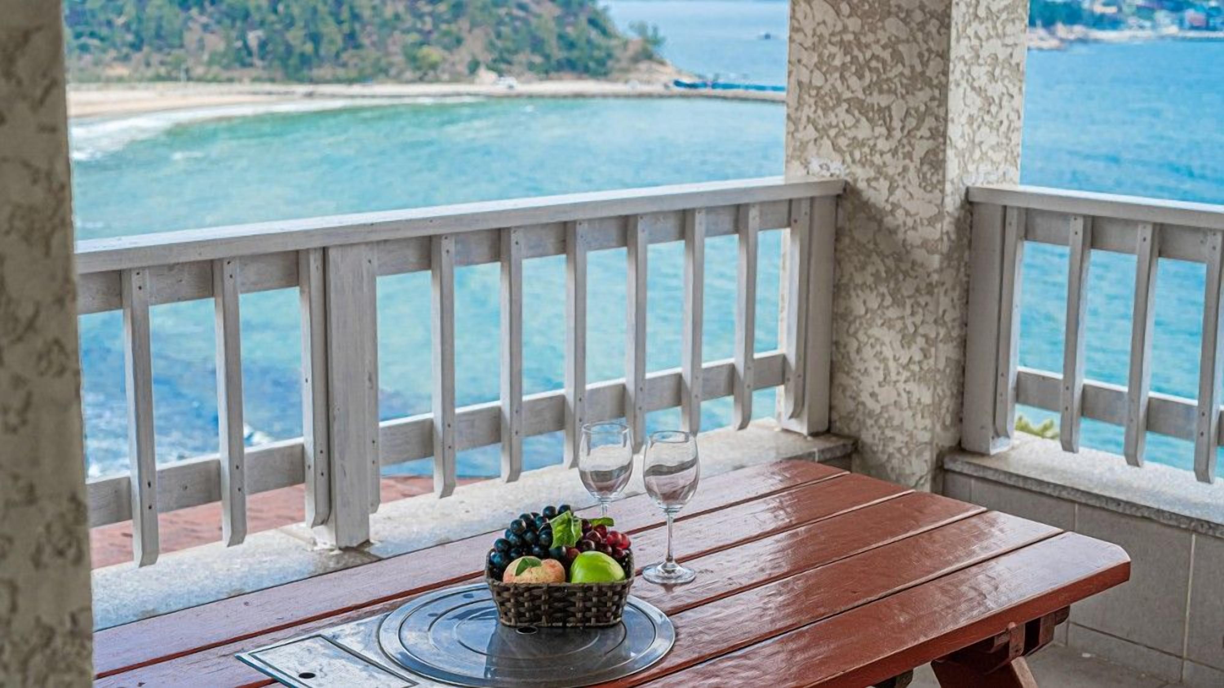A wooden table with a fruit basket and two wine glasses on a balcony overlooking a beach and the sea.