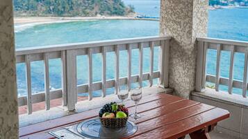 A wooden table with a fruit basket and two wine glasses on a balcony overlooking a beach and the sea.