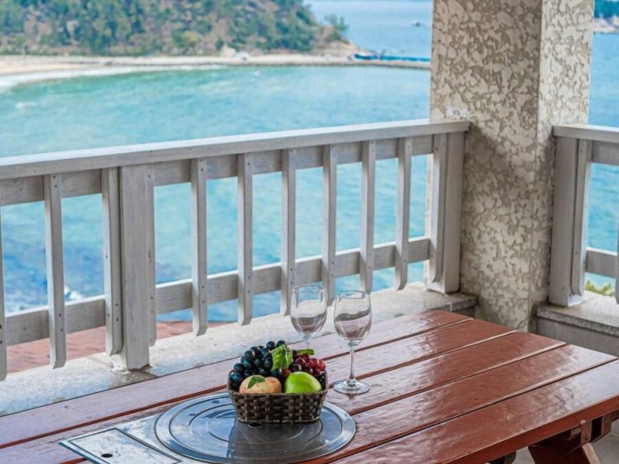 Una mesa de madera con una cesta de frutas y dos copas de vino en un balcón con vista a una playa y al mar.