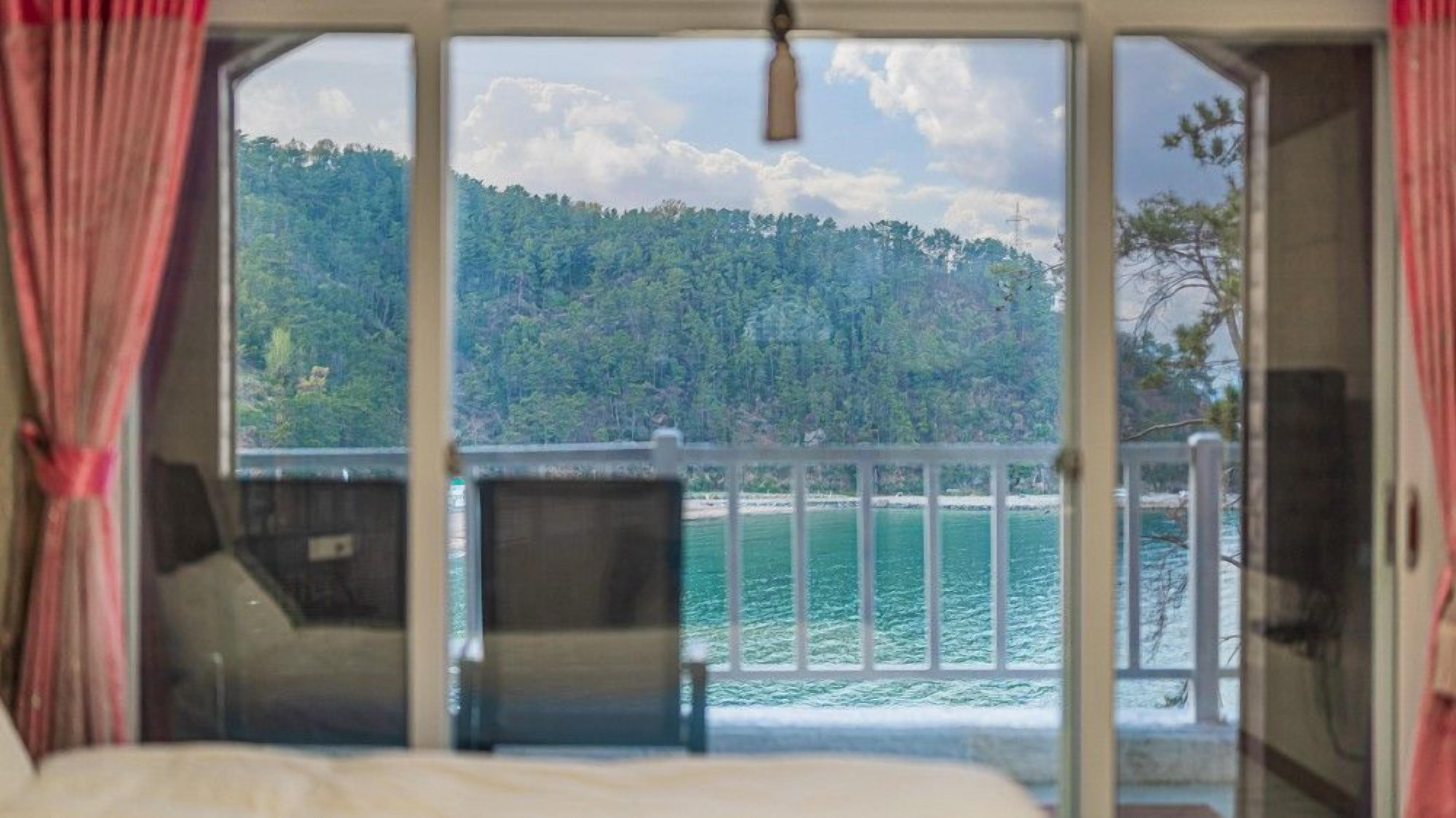 A room with a bed, a chair, and a view of a forested hillside and a body of water.
