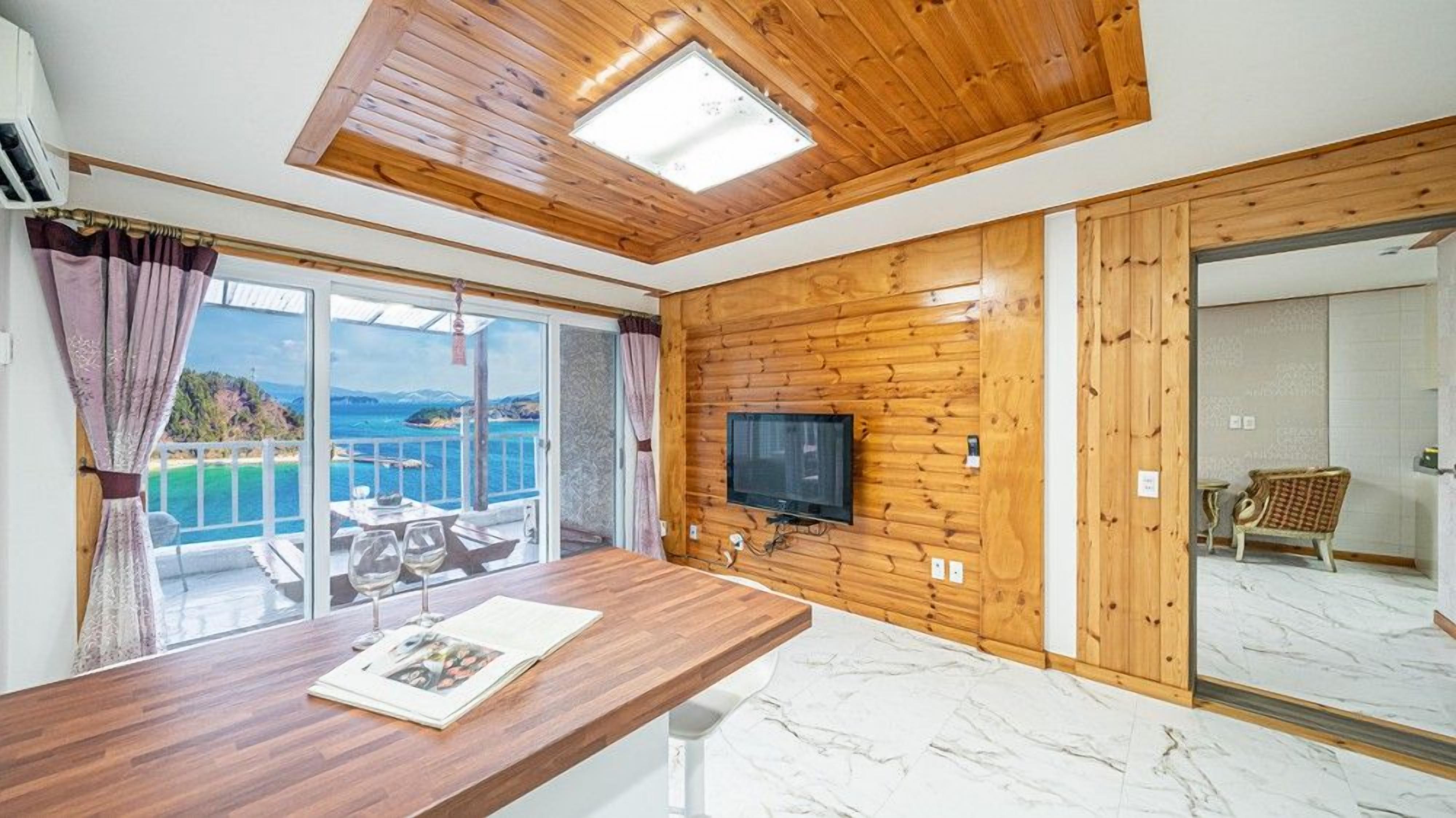 Une pièce avec un plafond en bois, un téléviseur fixé au mur, une table à manger avec un magazine, et une vue sur la mer par une porte vitrée.