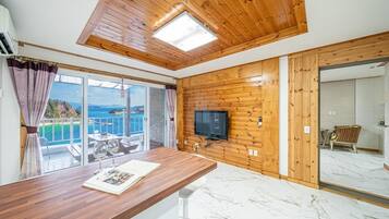 Une pièce avec un plafond en bois, un téléviseur fixé au mur, une table à manger avec un magazine, et une vue sur la mer par une porte vitrée.