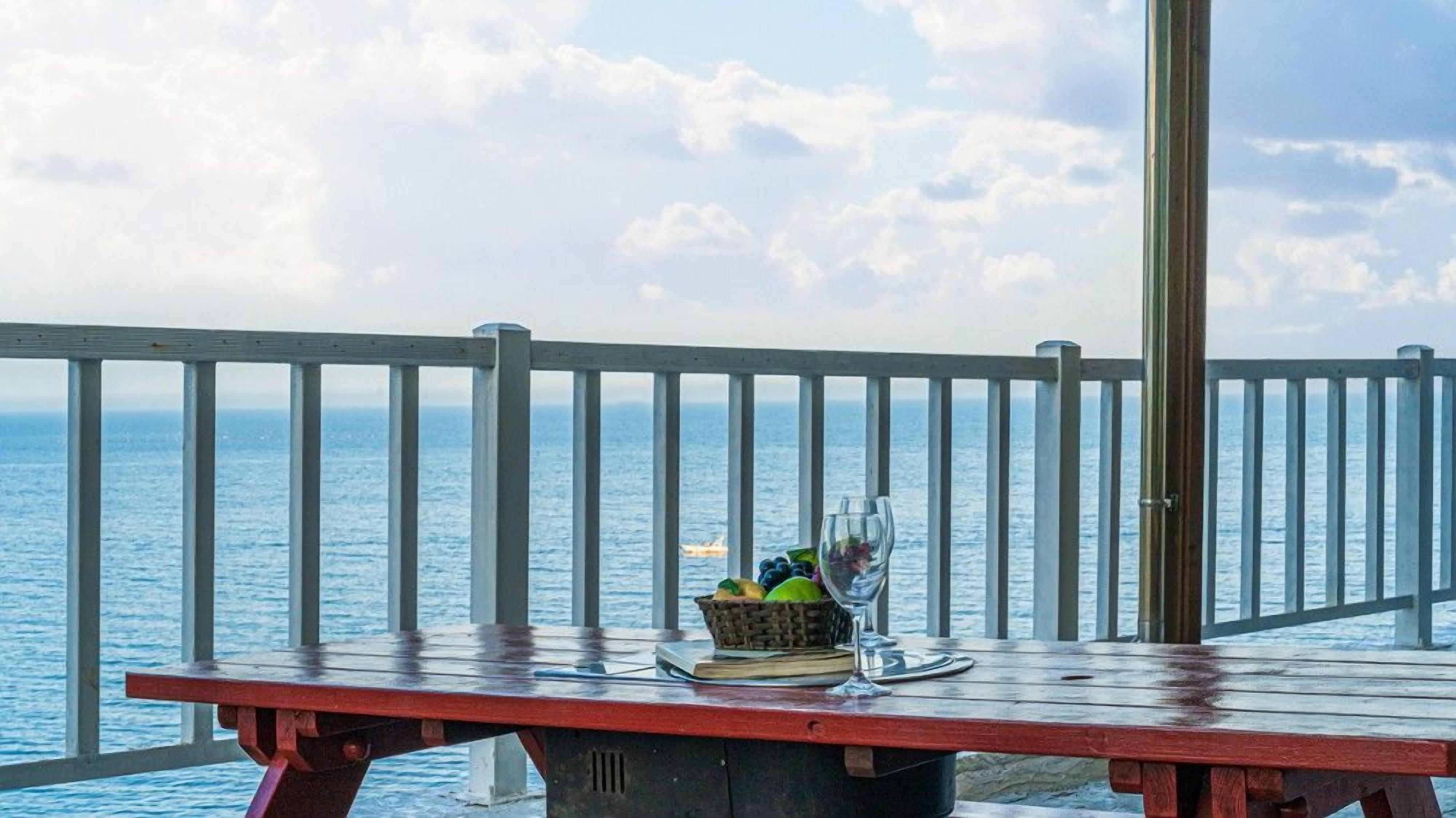A wooden table with a basket of fruit and a glass on it, overlooking the sea.