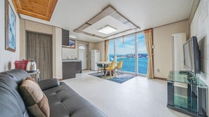 A modern living room with a sofa, a dining area with chairs, a kitchen, and a balcony with a view. - White O Pension (Geoje)