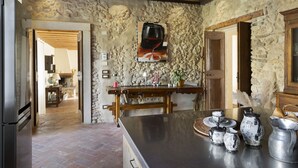 A rustic kitchen with stone walls, a wooden ceiling, and a stone fireplace. There is a wooden table with a painting of a wine glass, and various kitchen items on the counter. - CA’ DEL BUSO   (VERONA)