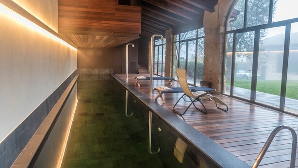 A modern indoor pool area with wooden decking, lounge chairs, and large windows offering views of the outdoors. - CA’ DEL BUSO   (VERONA)