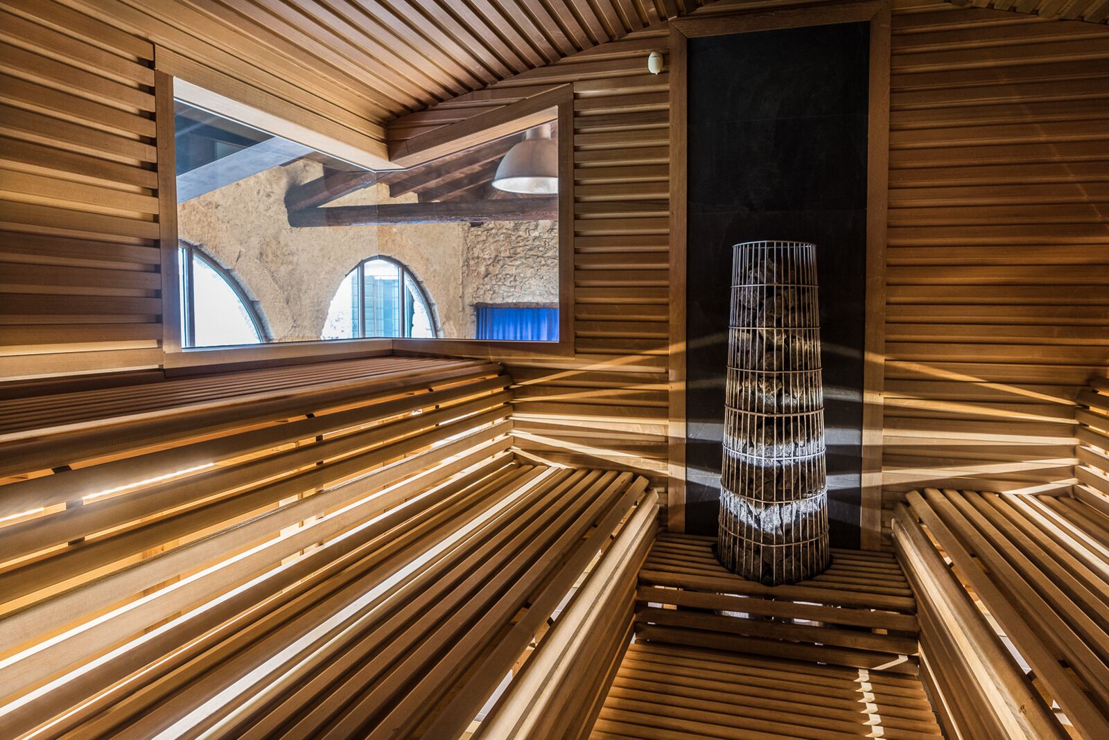 A modern interior with wooden slats on the walls and ceiling, a central cylindrical light fixture, and a view of an arched window.