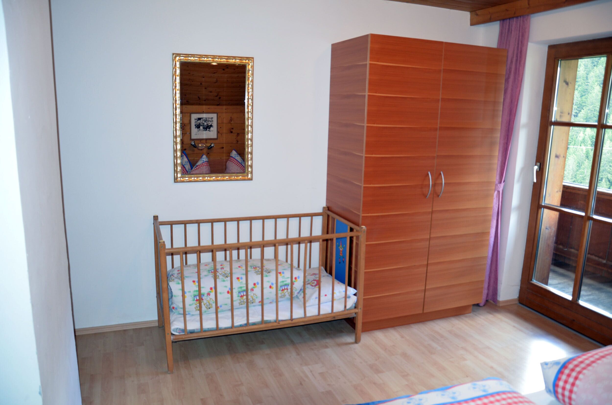 A room with a wooden crib, a wardrobe, and a window with a view of greenery.