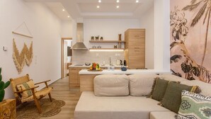 Una sala moderna con sofá y sillón, un área de cocina con mostrador y armarios, y una pared con un mural tropical.