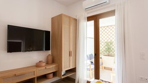 Una habitación moderna con un televisor de pantalla plana montado en la pared, un mueble de madera con espacio de almacenamiento, una mesita con un florero, y vistas a un patio exterior a través de una puerta de cristal.
