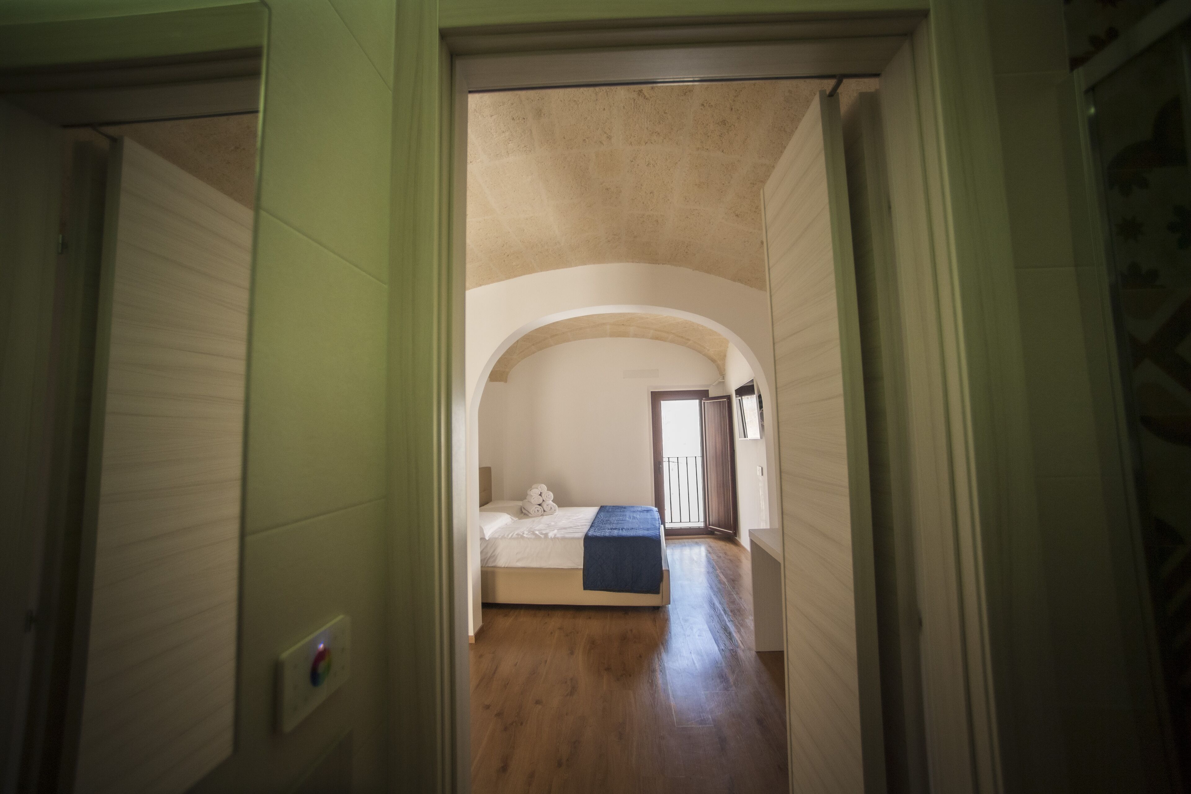 A narrow hallway leading to a bedroom with a single bed, a small window, and a door leading to another room.