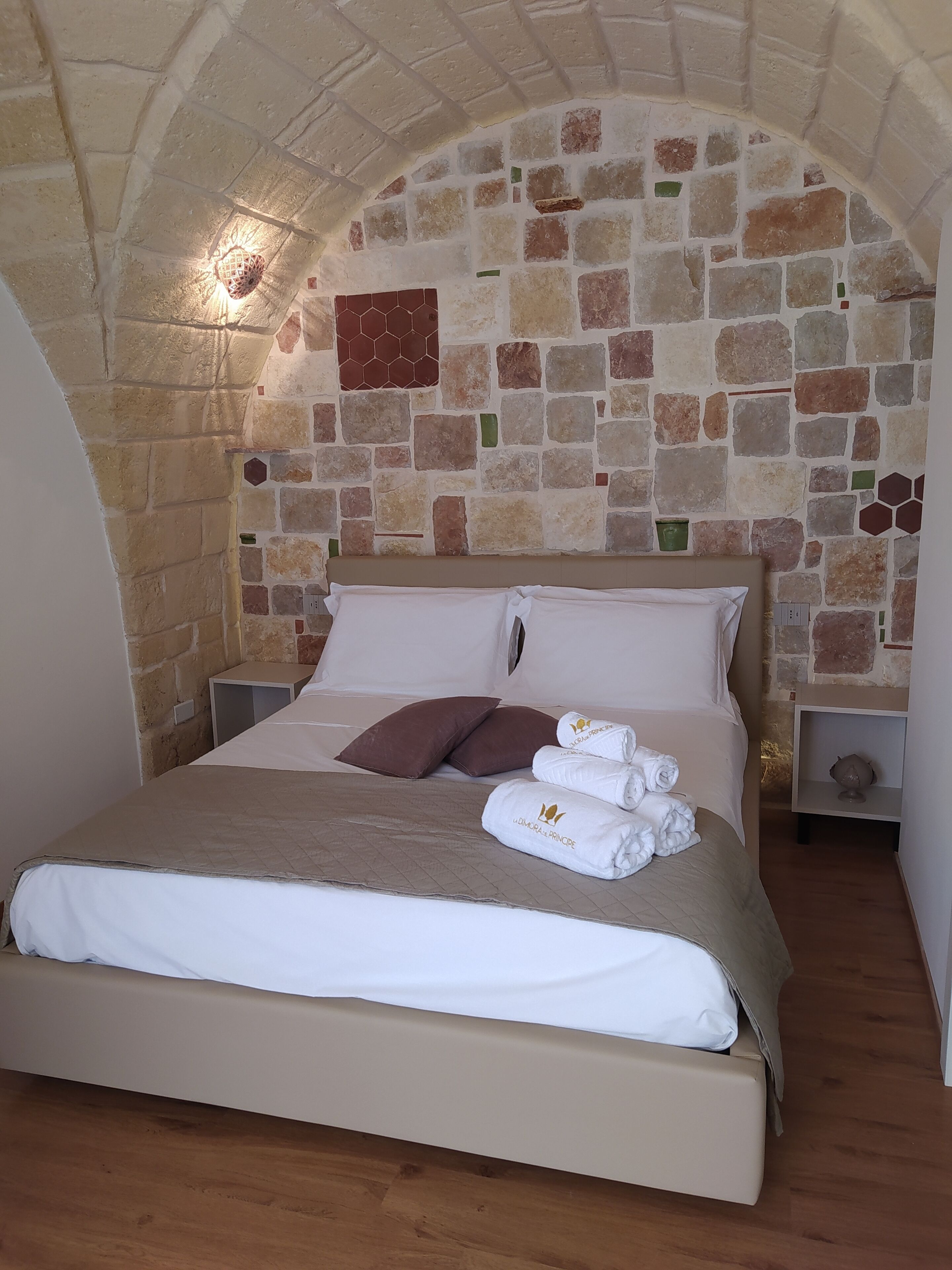A bed with a beige bedspread and pillows, a stone wall with a hexagonal tile pattern, and a small shelf with a decorative item.