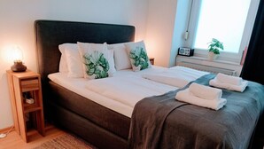 Een keurig opgemaakt bed met witte lakens en kussens met een bladmotief, een donker hoofdbord en een nachtkastje met een lamp.