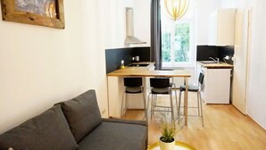 Een knus woongedeelte met een bank, een eettafel met stoelen, een kitchenette met een koelkast en spoelbak, en een raam met uitzicht op groen.