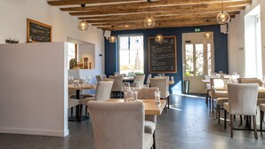 Interior de restaurante con vigas de madera a la vista, lámparas colgantes y un menú en la pizarra. Las mesas están dispuestas con sillas y el suelo está cubierto de baldosas.