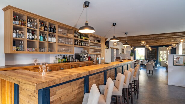 Una zona de bar con estanterías de madera llenas de botellas y vasos, una barra alargada y varios taburetes.