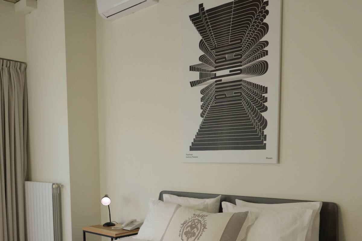 A neatly made bed with a beige bedspread, a nightstand with a lamp, and a wall-mounted air conditioner.