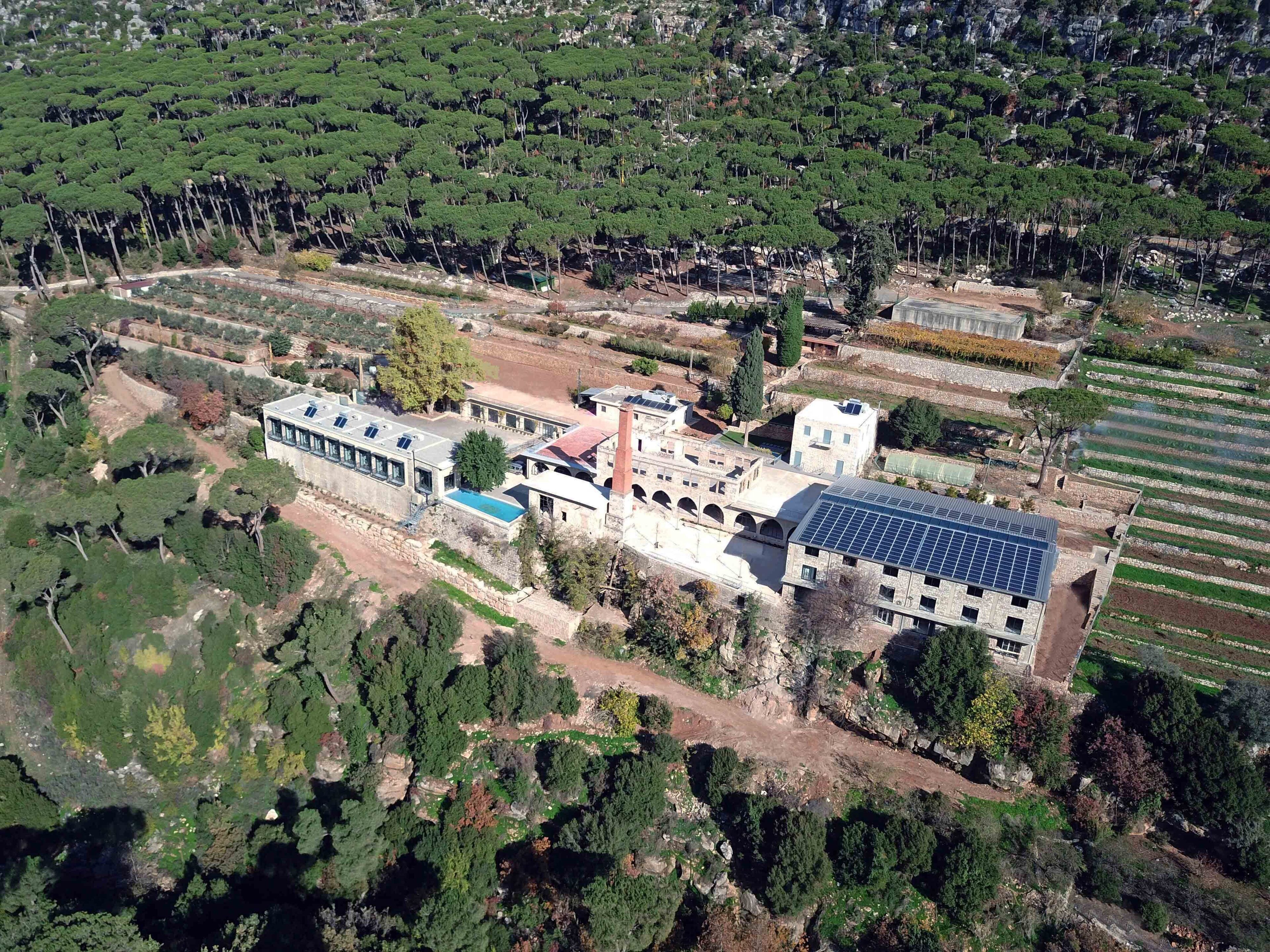 An aerial view of a building complex with multiple structures, including a large building with solar panels, surrounded by terraced fields and dense greenery.