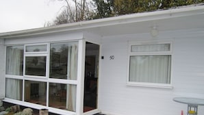 A white single-story house with a number '50' on the side, large windows, and a small patio area with a table and chair. - Upper Lake Chalet , Dog and Pet Friendly (Caernarfon)