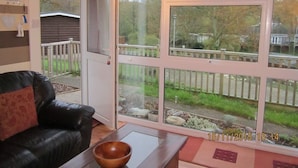 A living room with a black leather sofa, a wooden coffee table, and a large window with a view of a backyard and neighboring houses. - Upper Lake Chalet , Dog and Pet Friendly (Caernarfon)
