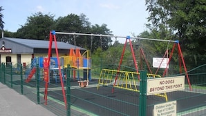 A fenced playground with a swing set and a slide, a 'No Dogs Beyond This Point' sign, and a 'Children Under The Age of 12 Must Be Accompanied By an Adult' sign. - Upper Lake Chalet , Dog and Pet Friendly (Caernarfon)