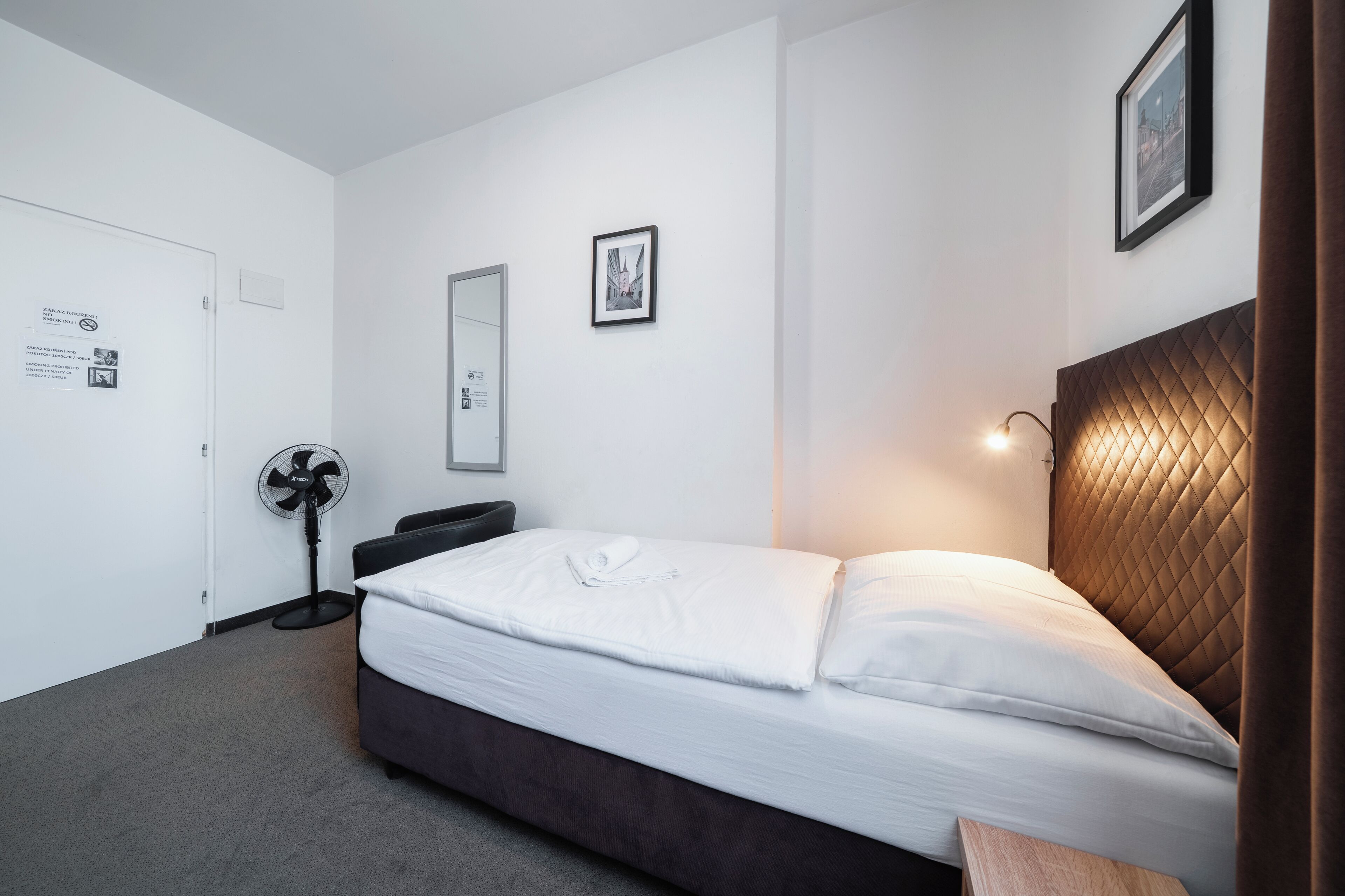 A hotel room with a bed, a bedside table, a wall-mounted fan, and a framed picture on the wall.
