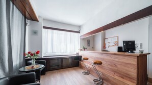 A modern hotel lobby with a wooden reception desk, two bar stools, a round table with a vase of flowers, and a large window with sheer curtains. - Penzion Homér Slaný (Slany)