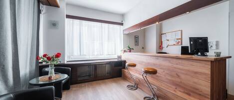 Un moderno vestíbulo de hotel con un mostrador de recepción de madera, dos taburetes de bar, una mesa redonda con un florero y un gran ventanal con cortinas translúcidas.