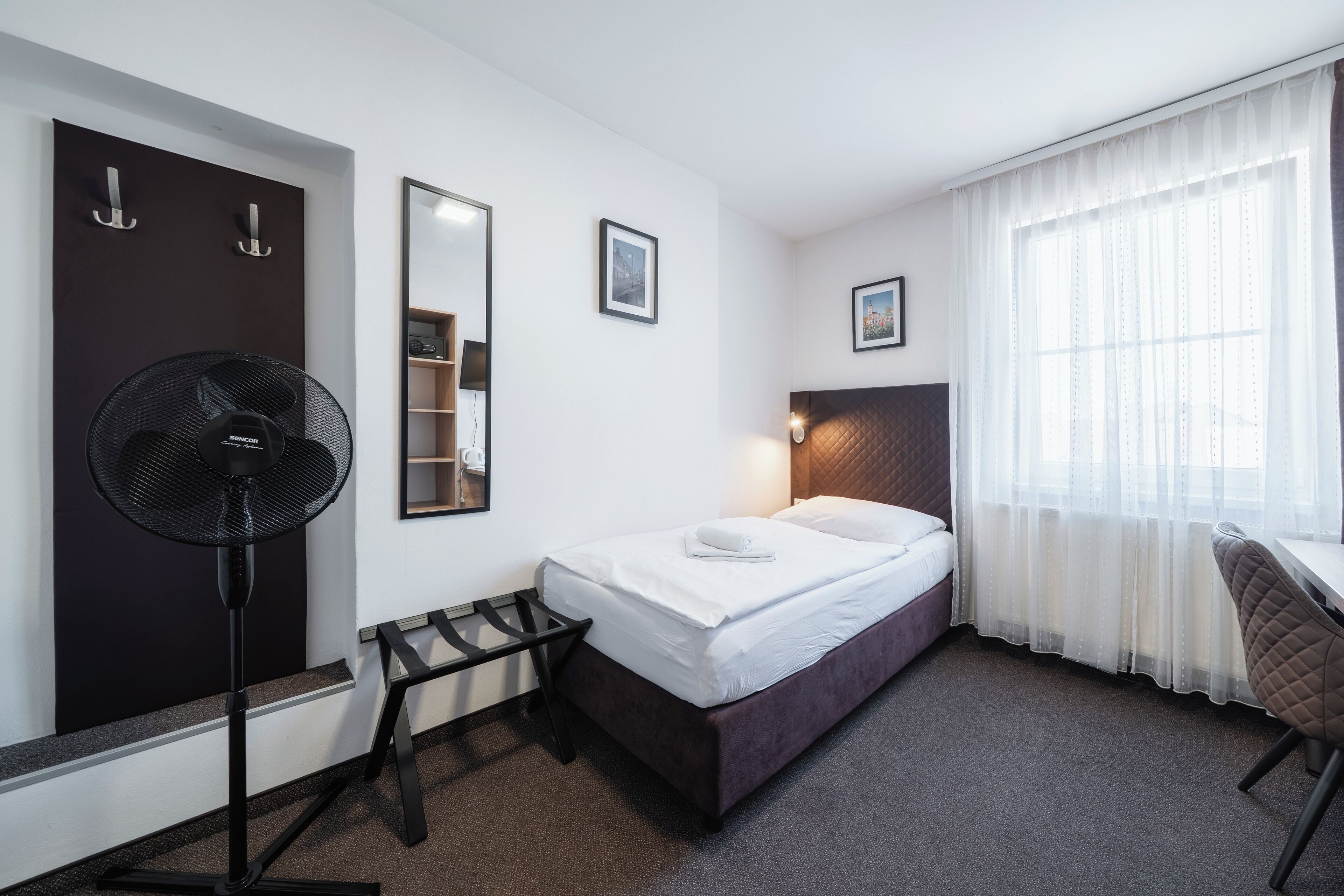 A hotel room with a bed, a desk, a chair, a standing fan, a wardrobe, and a window with curtains.