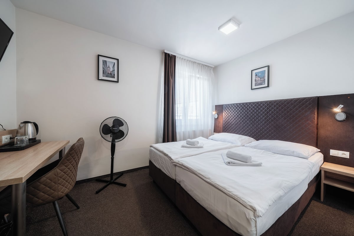 A hotel room with a bed, a desk with a kettle, a chair, a fan, and two framed pictures on the wall.